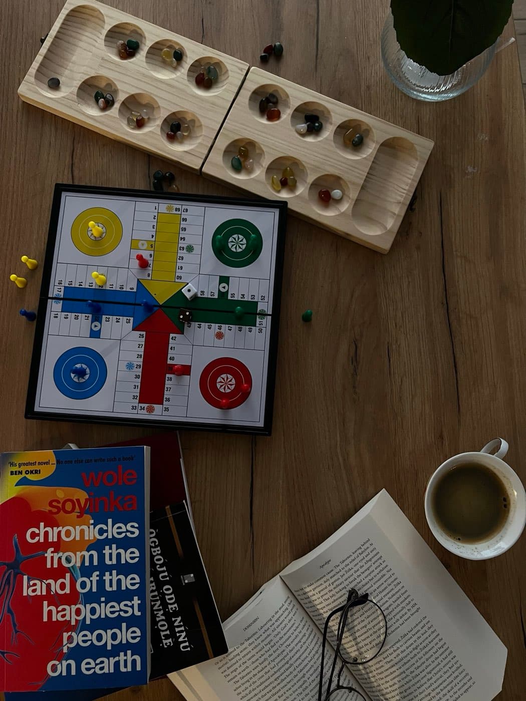 An ayo board, a ludo set, novels by Wole Soyinka and D.O. Fagunwa, and a cup of coffee laid out on a wooden table