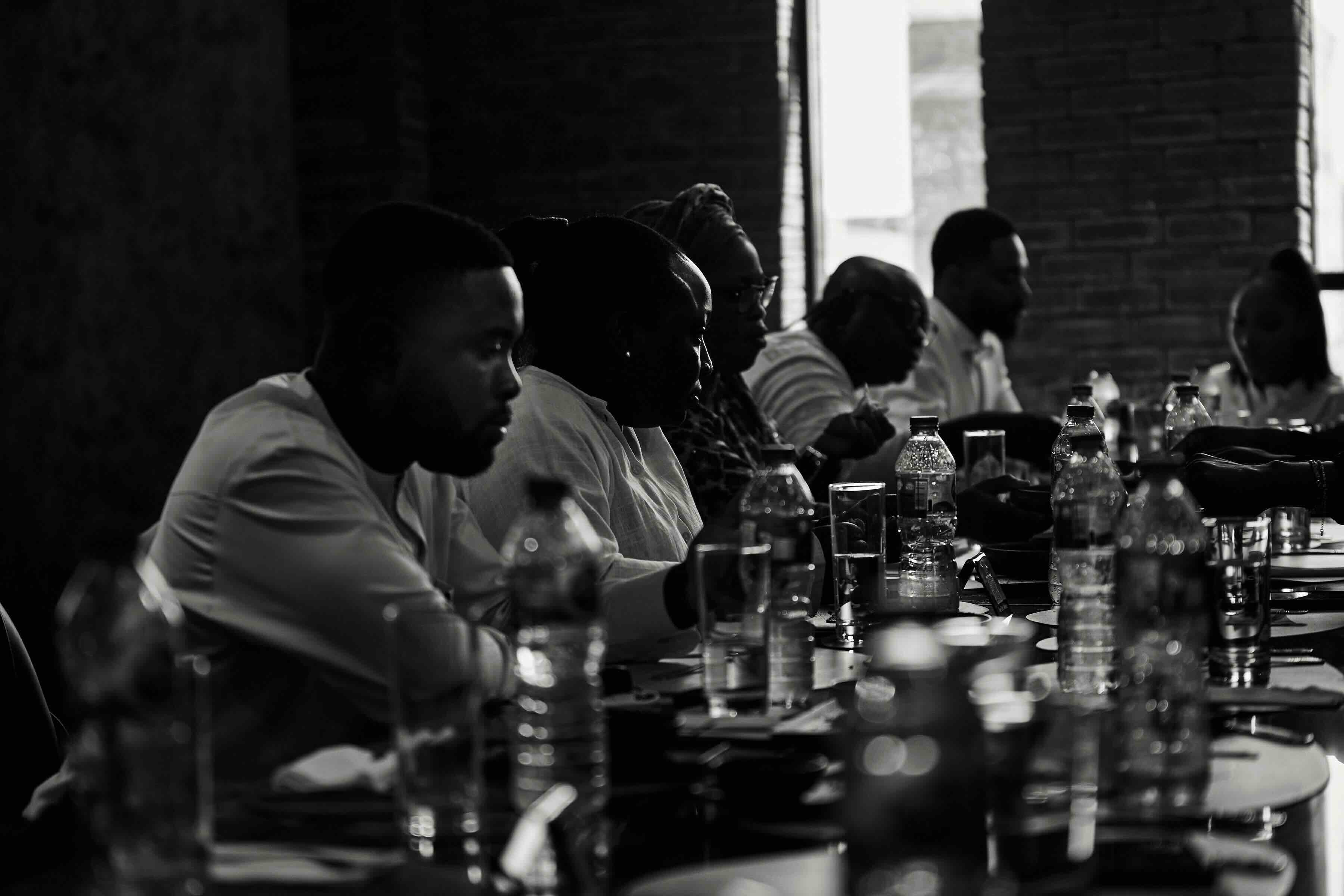 A long candlelit dinner table at a Good Yard gathering, guests in evening wear against deep shadow